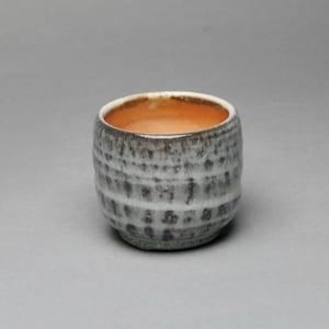 May include: A small, handmade ceramic cup with a textured, gray and white exterior. The interior of the cup is a warm orange color. The cup has a rounded shape and a slightly uneven rim, suggesting a handcrafted origin.