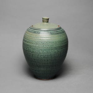 May include: A handcrafted, green ceramic jar with a lid. The jar has a rounded shape with horizontal textured lines. The lid is a lighter shade of green and has a small knob on top. The jar is a decorative storage container.