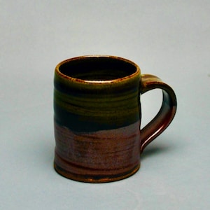 May include: A handmade ceramic mug with a cylindrical shape and a curved handle. The mug features a color gradient, transitioning from brown at the base to dark green and black in the middle, and a brown rim. The mug is suitable for hot or cold beverages.