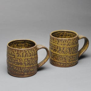 May include: Two ceramic mugs featuring a textured, mustard-yellow and brown crackle glaze pattern. Each mug has a cylindrical body, a flat base, and a curved handle on the side. The interior and exterior surfaces display a consistent, raised, web-like pattern.
