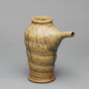 May include: A handmade ceramic pitcher with a unique design. The pitcher features a light yellow base color with brown and gray accents. It has a spout extending from the side and a textured, organic form. The pottery is suitable for decorative use.