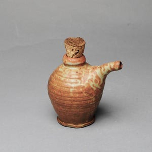 May include: A handmade ceramic pitcher with a cork stopper. The pitcher is a warm, earthy orange with lighter tan and green accents. It has a rounded body, a small spout, and a textured surface. The cork is a light brown color.