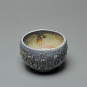 May include: A ceramic bowl with a textured, dark blue exterior and a light yellow interior. The bowl has a rounded shape and a slightly rough surface. The interior has a mottled appearance with hints of orange and brown.