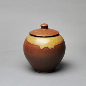 May include: A ceramic jar with a lid. The jar is a rounded shape with a dark brown base, transitioning to a yellow-tan band, and topped with a brown lid. The lid has a small knob. The jar is likely for storage or decoration.
