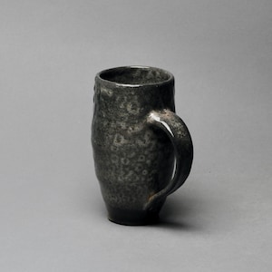 May include: A dark gray ceramic mug with a speckled glaze and a handle. The mug is slightly taller than it is wide.