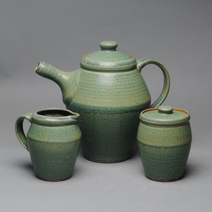 May include: A green ceramic teapot, creamer, and sugar bowl set. The teapot has a lid and a spout. The creamer and sugar bowl have lids.