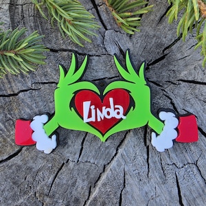May include: A decorative ornament featuring two bright green hands forming a heart shape around a red heart. The name Linda is written in white script across the centre of the red heart. The hands have white cuffs and red sleeves at the ends.