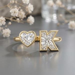 Op de afbeelding: Een gouden ring met een open ontwerp, voorzien van een hartvormige heldere edelsteen aan de ene kant en een heldere edelsteen in de vorm van de letter K aan de andere kant. De ringband is van gepolijst goud en beide stenen zijn gevat in goud.