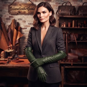 Peut inclure: Une paire de longs gants en cuir vert olive foncé avec des détails cousus sur le dos des mains. Les gants dépassent les poignets et atteignent les coudes, portés avec un blazer gris anthracite foncé dans un atelier de maroquinerie.