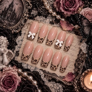 Peut inclure: Un ensemble de dix faux ongles avec une base nude et des pointes françaises à motif léopard marron et noir. Deux des ongles sont ornés de petits nœuds blancs en relief placés à la jonction entre la base nude et le motif léopard.