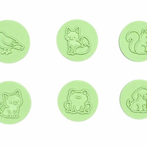 Puede incluir: Un conjunto de seis sellos circulares de color verde claro, cada uno con el contorno en relieve de un animal. Los diseños incluyen un pájaro, un zorro, una ardilla, un gato, una rana y un perro, todos en un estilo de línea simple.
