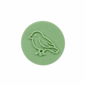 May include: A circular light green stamp featuring a raised outline of a small bird in profile. The bird design is centered on the flat disc, showing a simple silhouette with a beak, eye, wing, and tail feathers, all rendered in a monochromatic tone.