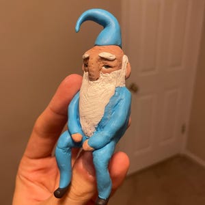 Puede incluir: Una pequeña figura esculpida a mano de un gnomo sentado que viste un conjunto azul brillante y un sombrero puntiagudo a juego. El gnomo tiene una barba blanca texturizada, cejas blancas, cara color canela y pequeños zapatos marrón oscuro.
