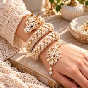 Puede incluir: Una pulsera de dragón tejida a ganchillo en color crema con una cabeza de dragón detallada, acentos dorados y ojos negros. El cuerpo está enrollado con perlas blancas incrustadas en la textura, terminando en una cola con forma de hoja.