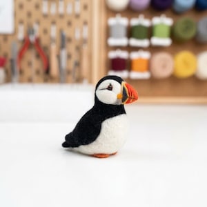 May include: A needle felted puffin figurine featuring a black back, white chest, and a distinctive beak with orange, yellow, and black stripes. The small bird sculpture has a rounded body, a dark eye, and small orange feet, crafted with a soft, textured wool finish.
