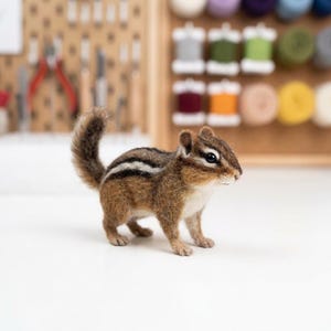 May include: A needle-felted chipmunk figurine featuring a textured brown coat with distinct black and white stripes running along its back. The small animal has a bushy tail, dark eyes, and a light-colored underside, standing in a realistic pose.