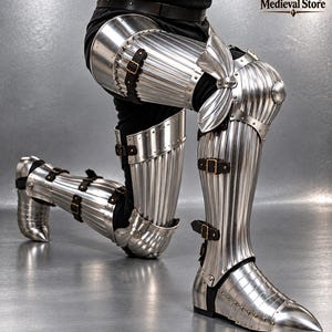 May include: A set of polished steel leg armour featuring vertical fluting, articulated knee cops with side wings, and pointed sabatons. The armour is secured with dark brown leather straps and brass buckles, covering the thigh, shin, and foot.