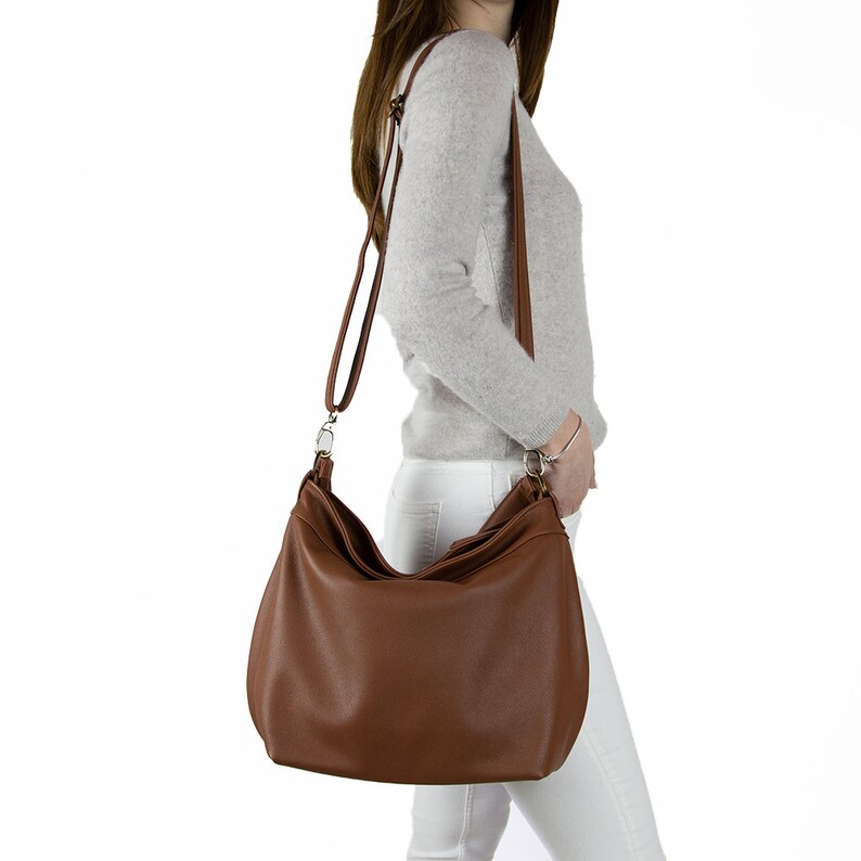 May include: A brown hobo-style shoulder bag with a smooth finish, featuring a long adjustable strap attached with silver-toned metal hardware. The bag has a slouchy, rounded silhouette and hangs at the hip, providing a spacious main compartment.