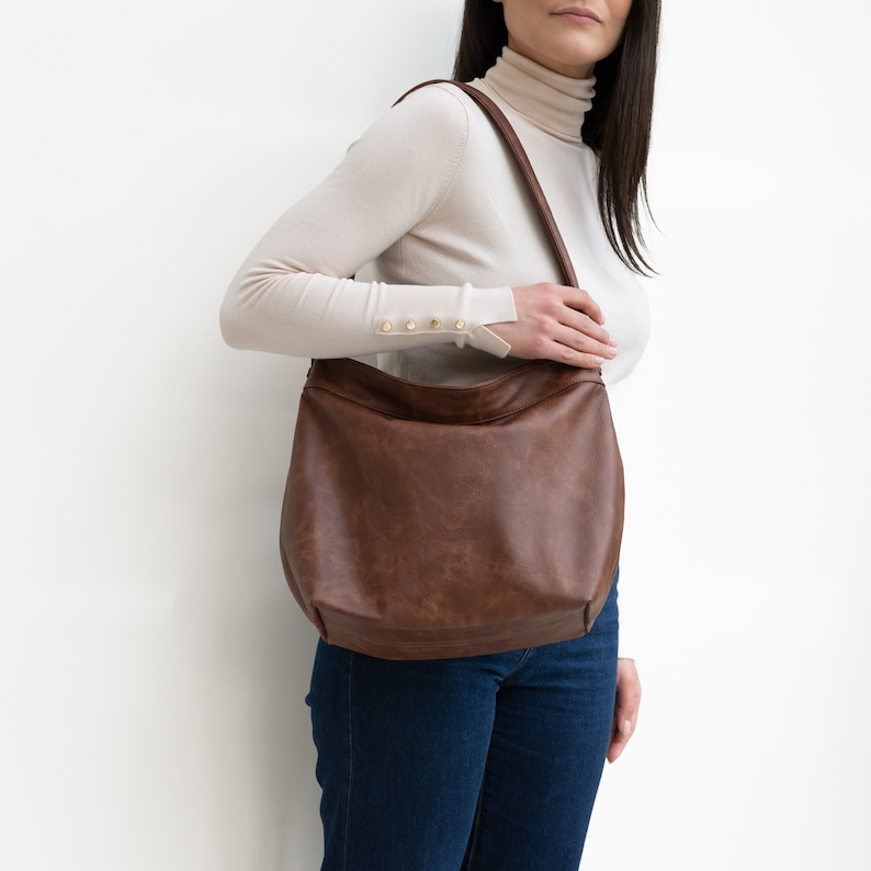 May include: A brown leather tote bag with a long strap. The bag is worn by a person wearing a white turtleneck sweater and blue jeans.