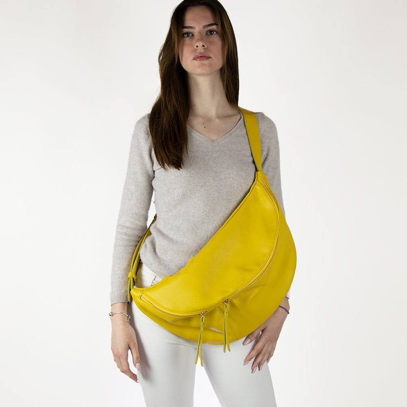 May include: A bright yellow leather crossbody bag with a curved shape and two zipper pulls. The bag is worn by a person with long brown hair and a light grey sweater.
