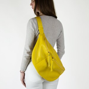May include: A bright yellow leather fanny pack with a gold zipper and a long strap. The bag is worn on the shoulder of a person wearing a grey sweater and white pants.