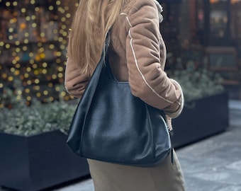 May include: A woman wearing a brown suede jacket with a white trim carries a black leather tote bag with a slouchy design.
