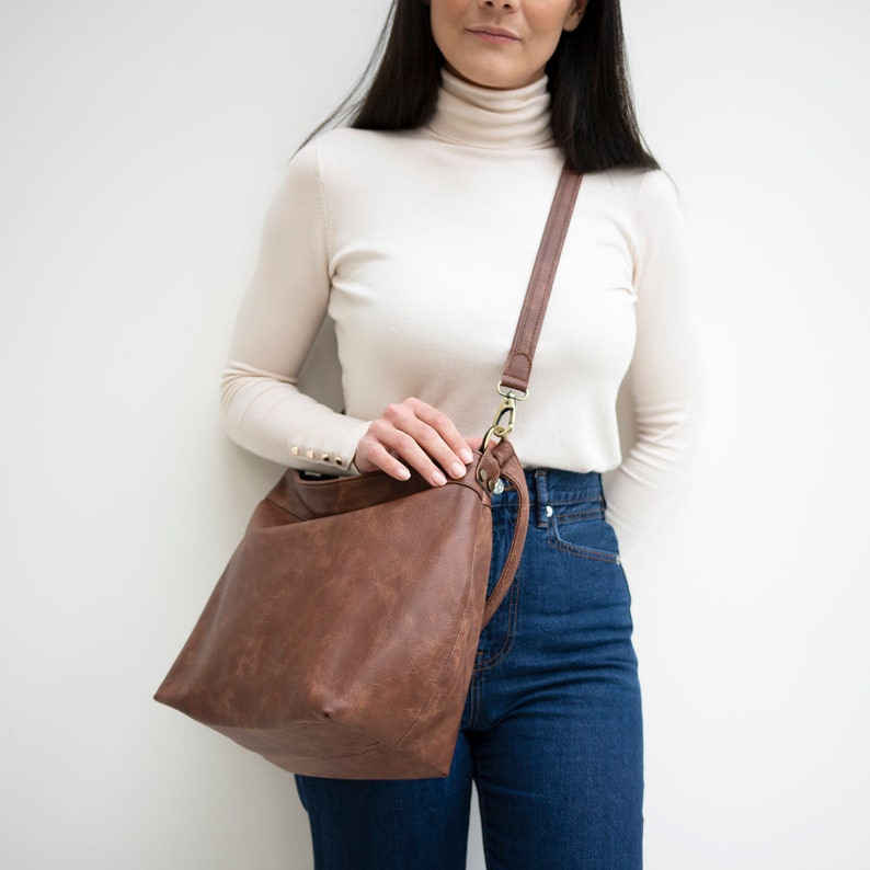 May include: A brown leather shoulder bag with a long strap. The bag is worn by a person wearing a white turtleneck sweater and blue jeans.