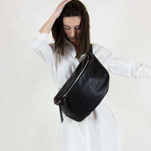 Könnte beinhalten: Eine schwarze Leder-Bauchtasche mit einem silbernen Reißverschluss. Die Tasche wird über der Schulter getragen.