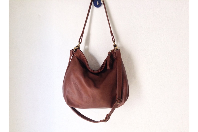 May include: A medium-sized brown leather hobo bag with a slouchy silhouette, featuring a long adjustable shoulder strap, antique brass-toned metal hardware, and a top zipper closure. The bag has a textured surface and hangs from a small blue hook.