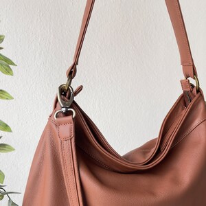 May include: A terracotta-colored hobo bag featuring a smooth, textured surface, a top zipper closure, and a matching shoulder strap attached with antique brass-toned metal swivel clasps and D-rings, showcasing a slouchy, unstructured design.