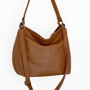 May include: A medium-sized hobo-style shoulder bag in a solid tan brown color. The bag features a smooth exterior, a single main compartment with a zipper closure, and an adjustable shoulder strap attached with antique brass-toned metal hardware.