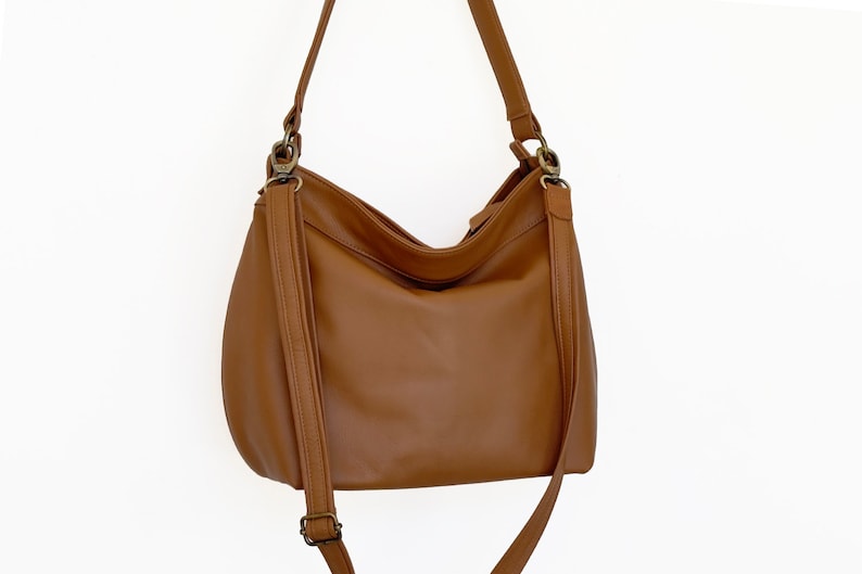 May include: A medium-sized hobo-style shoulder bag in a solid tan brown color. The bag features a smooth exterior, a single main compartment with a zipper closure, and an adjustable shoulder strap attached with antique brass-toned metal hardware.