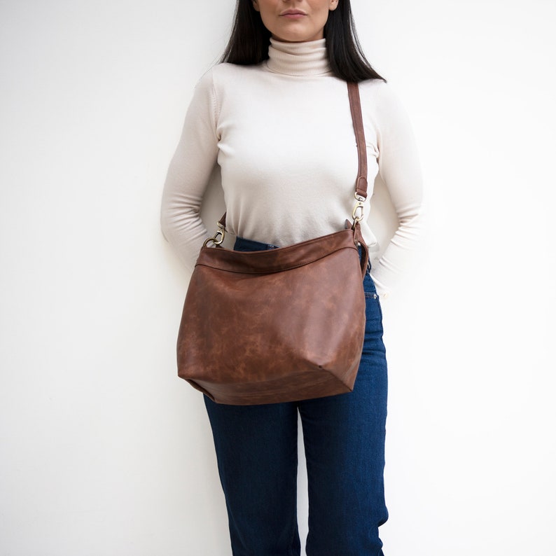 May include: A brown leather shoulder bag with a long strap. The bag is worn by a person wearing a white turtleneck sweater and blue jeans.