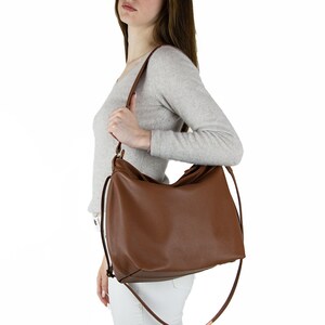 May include: A medium-sized, solid brown hobo-style shoulder bag with a smooth, matte finish. The bag features a wide, curved main compartment, a single top handle, and a long, thin adjustable shoulder strap attached to the sides with gold-tone hardware.