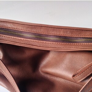 May include: A brown leather handbag featuring a textured surface with a visible top zipper closure. The bag has a dark brown fabric lining along the zipper track and includes a matching brown leather strap attached to the side of the bag.