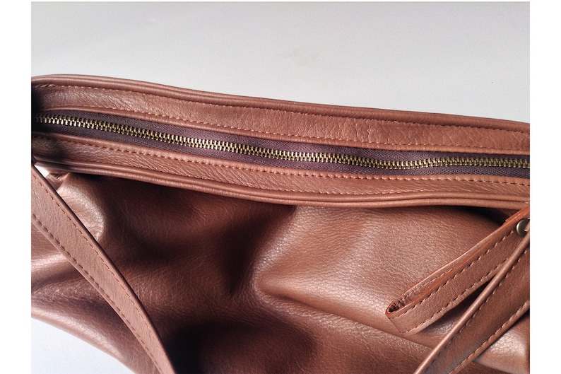 May include: A brown leather handbag featuring a textured surface with a visible top zipper closure. The bag has a dark brown fabric lining along the zipper track and includes a matching brown leather strap attached to the side of the bag.