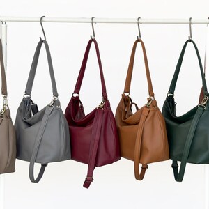 May include: Six hobo-style shoulder bags hanging on a white metal rack. The bags are available in taupe, gray, burgundy, tan, dark green, and brown. Each bag features a single shoulder strap, metallic hardware, and a smooth, solid-colored exterior.