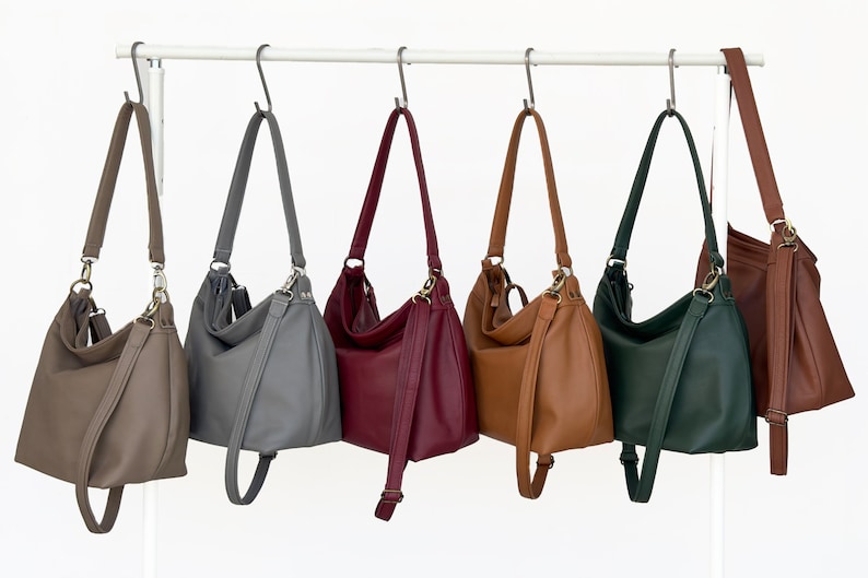 May include: Six hobo-style shoulder bags hanging on a white metal rack. The bags are available in taupe, gray, burgundy, tan, dark green, and brown. Each bag features a single shoulder strap, metallic hardware, and a smooth, solid-colored exterior.
