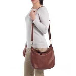 May include: A medium-sized brown leather hobo bag featuring a smooth texture, a single main compartment with a top zipper closure, and an adjustable shoulder strap attached with gold-toned metal hardware clips on both sides of the bag.
