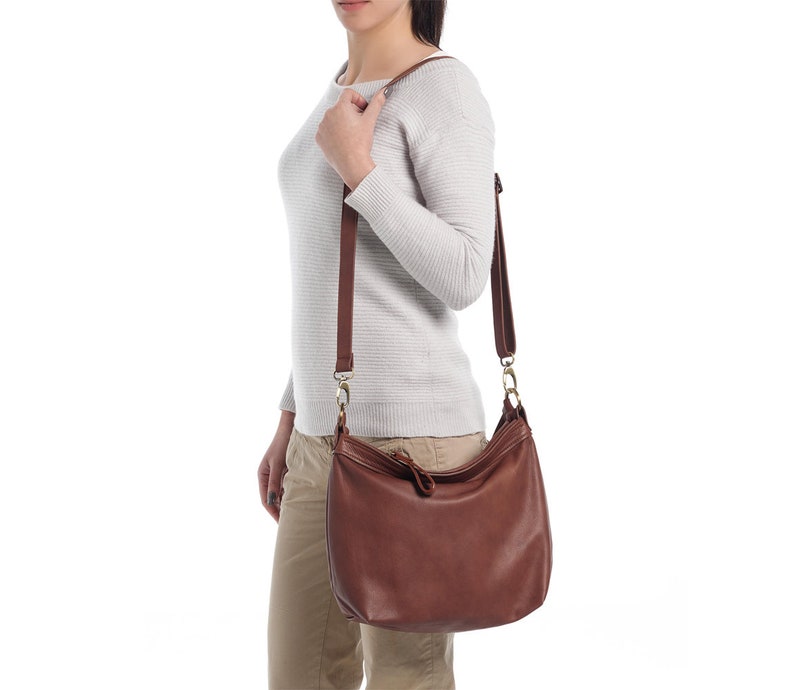 May include: A medium-sized brown leather hobo bag featuring a smooth texture, a single main compartment with a top zipper closure, and an adjustable shoulder strap attached with gold-toned metal hardware clips on both sides of the bag.
