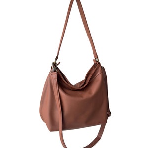 May include: A solid terracotta-colored hobo bag featuring a slouchy silhouette, a single main compartment with a zipper closure, and an adjustable shoulder strap attached with metallic hardware clips on both sides of the bag's upper opening.