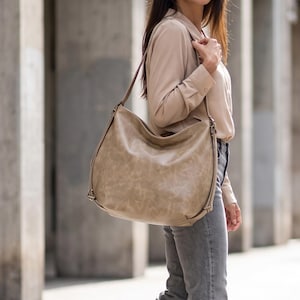 May include: A large, slouchy hobo-style shoulder bag in a muted taupe or beige leather with a subtle distressed texture. The bag features a single wide shoulder strap, visible side hardware attachments, and a spacious, unstructured main compartment.