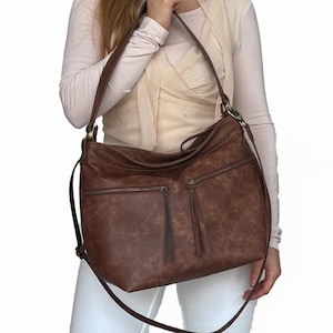 Könnte beinhalten: Eine braune Ledertasche im Satchel-Stil mit einem langen, verstellbaren Trageriemen. Die Tasche hat mehrere Reißverschlussfächer und ein Quasten-Detail.