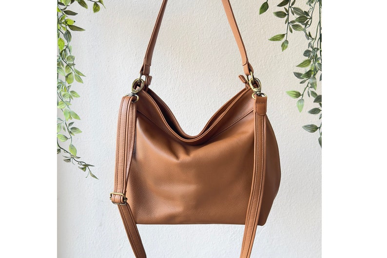 May include: A tan hobo-style shoulder bag with a smooth, matte finish. The bag features a wide main compartment, a single top handle, and an adjustable crossbody strap attached with antique brass-toned metal hardware clips and side rings.