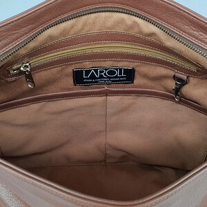 May include: The interior of a brown leather bag featuring a tan fabric lining, a top zipper closure, and an internal zippered pocket. A black rectangular label is stitched to the lining with the white text LAROLL STYLISH & FUNCTIONAL LEATHER BAGS.