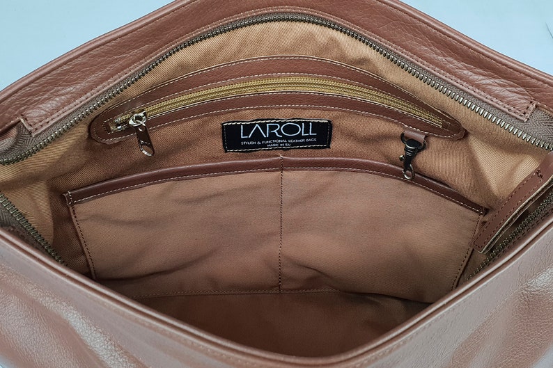 May include: The interior of a brown leather bag featuring a tan fabric lining, a top zipper closure, and an internal zippered pocket. A black rectangular label is stitched to the lining with the white text LAROLL STYLISH & FUNCTIONAL LEATHER BAGS.