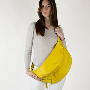 May include: A bright yellow leather crossbody bag with a zipper closure. The bag is worn by a person with long brown hair and a light gray sweater.