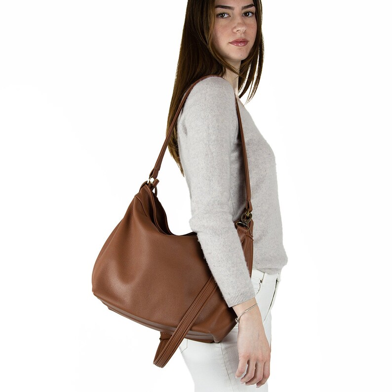 May include: A medium-sized, slouchy hobo-style shoulder bag in a solid brown color. The bag features a smooth, matte texture, a single wide shoulder strap, and a dangling strap detail on the side, constructed with visible stitching along the edges.