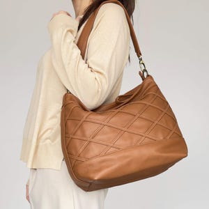 Könnte beinhalten: Hellbraune Leder-Umhängetasche mit einem gesteppten Rautenmuster. Die Tasche hat einen langen Riemen und einen goldfarbenen Verschluss. Die Tasche wird über der Schulter getragen.