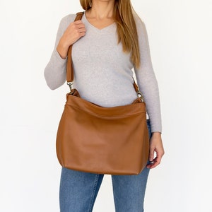 May include: A medium-sized, smooth tan leather shoulder bag with a wide, adjustable strap and antique brass-toned metal hardware. The bag features a soft, slouchy silhouette with a top zipper closure and a clean, minimalist exterior design.